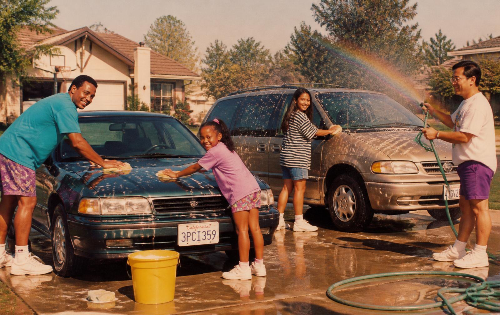 A family and neighbors hand-washing cars in a sunny 1990s driveway with sponges, a yellow bucket, and a garden hose